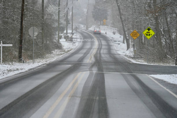 Fairfax, Virginia, USA - January 15, 2024: Falling snow quickly covers a road through a wooded neighborhood as a solitary car carefully ascends a slight hill during the first significant snowstorm of the new year.