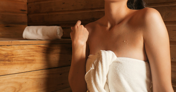 Young woman in towel relaxing in wooden sauna at spa. Asian woman in bathrobe doing body treatment in sauna.