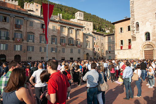 Gubbio Umbria, Italy-May 15 2011; Crowd facing away gather in European town square as sun lowers and shadows lengthen in late afternoon.