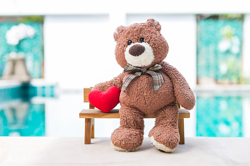 Brown teddy bear sitting on wooden bench over blurred swimming pool background, outdoor day light, little teddy bear holding red heart sitting on wooden chair, valentine concept background idea