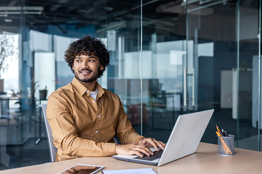 Indian young smiling man working in office at desk with laptop and looking confidently to side.