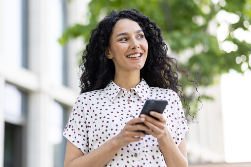 A young beautiful Latin American woman with curly hair walks through the city with a phone in her hands, a woman uses an application on a smartphone, browses Internet pages and social networks.