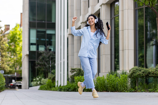 Young joyful beautiful woman dancing and singing in headphones while walking in the city, Hispanic woman with curly hair uses an application on the phone, to listen to music online.