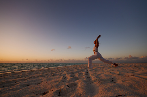 Carefree woman exercising Pilates with her arms raised during summer sunset on the beach. Copy space.