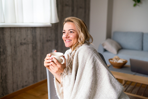 Happy woman whit a blanket sitting on a chair and holding cup of coffe at home