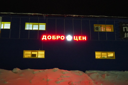 Petrozavodsk, Karelia, Russia, January 11, 2024: building - hangar of the Dobrotsen store, at night in winter. Red-yellow glow-in-the-dark letters and words on the facade. Saving money. Low prices.