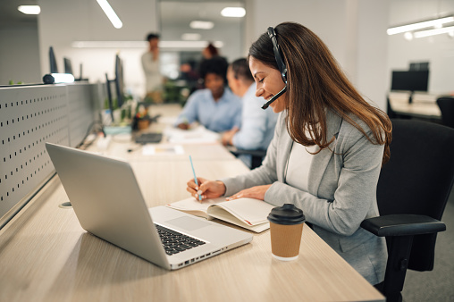 Portrait of a customer support operator sitting at call center at the office with hands free headset and giving desk solutions to a customer on the call. A friendly agent solving problems on a call.