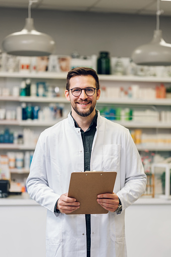 A young pharmacy intern doing inventory at the pharmacy shop as a part of their internship. He wears glasses and a white pharmacy coat. He is in front of a counter. Blurry background with shelves.