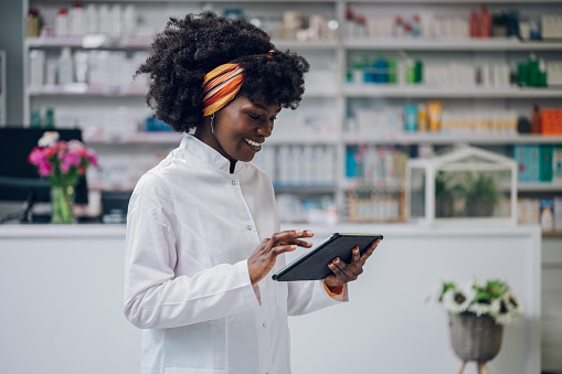 Portrait of a young African-American druggist standing in a drugstore with a tablet in her hands and smiling at it while scrolling. An apothecary worker is selling drugs online.