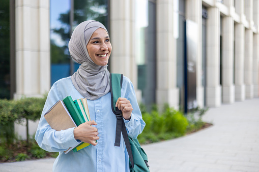 A cheerful young Arab woman in a hijab holding notebooks, walking by an office, exuding confidence and professionalism.