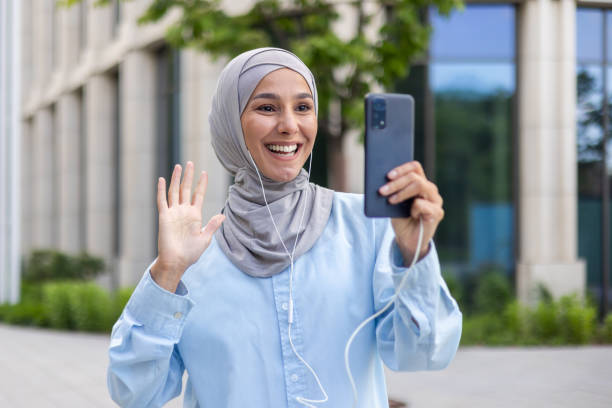 Muslim woman in hijab using smartphone, greeting with a wave, urban background