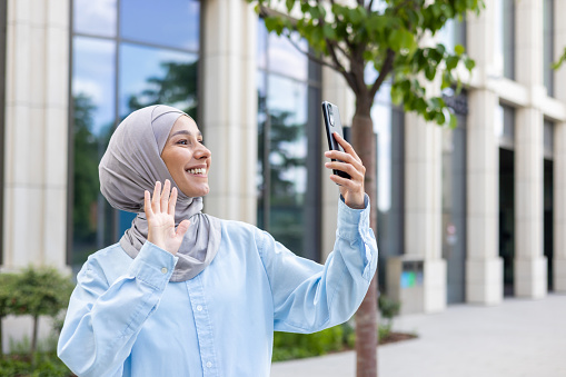 Beautiful Arab businesswoman in hijab using mobile phone for video call, happily waving at the screen outdoors.