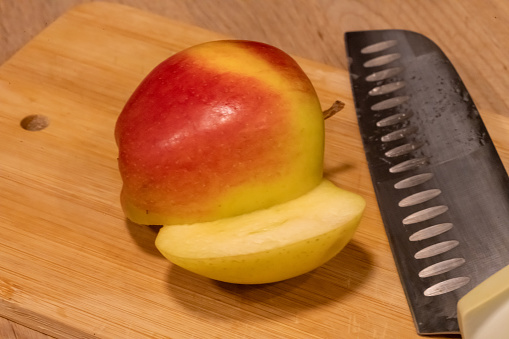An English Cox apple sliced in half sitting on a wooden chopping board with its bright red skin markings reflecting the artificial light with a chopping knife laid by its side