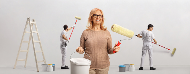 Happy woman with a paint roller and a bucket with workers painting a wall isolated on white background