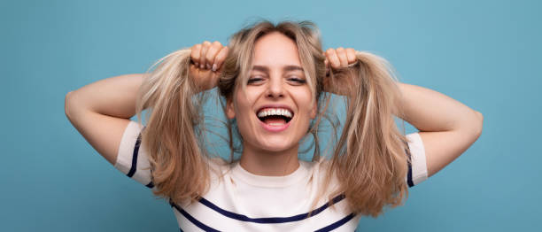 cheerful blond young woman holds her hair in her hands and laughs cutely on a blue background - mid adult women human skin neat good condition photos et images de collection