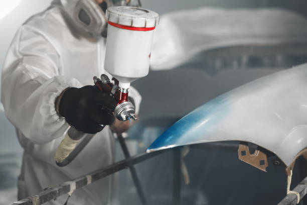 istock Car paint worker spraying blue paint to car body element using spray gun in vehicle workshop chamber. Complex restoration of automobile body. 1969514806