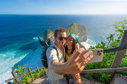 Family vacation lifestyle. Happy multiracial couple stand at viewpoint, looking at the beautiful beach under high cliff. Travel destination in Bali. Popular place to visit on Nusa Penida island.