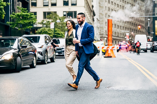 Full length view of male and female colleagues in 20s and 30s running across city street with takeaway coffee, summertime.