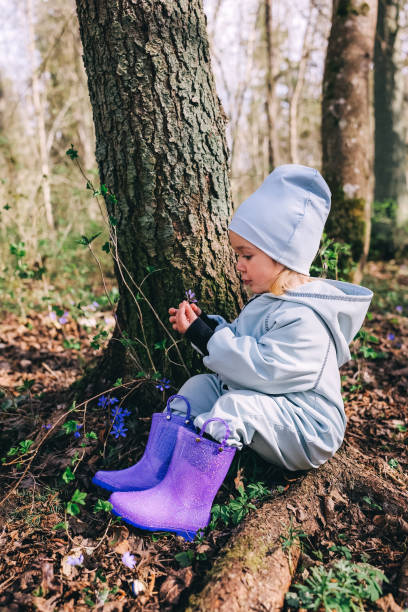 Little girl in violet boots and blue overall sitting under the tree in spring forest Little girl in violet boots and blue overall holding violet flowers in hands sitting under the tree in spring forest covered with violet flowers Hepatica nobilis holding child flower april stock pictures, royalty-free photos & images