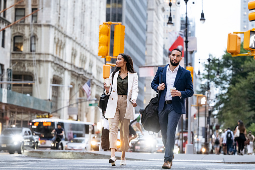 Young multiracial woman and mid adult Hispanic man in summer attire, carrying shoulder bags and coffee, crossing the street.