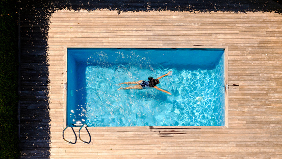 Drone view full length of unrecognizable woman with long wet dark hair floating in outdoor swimming pool on sunny day in tropical resort