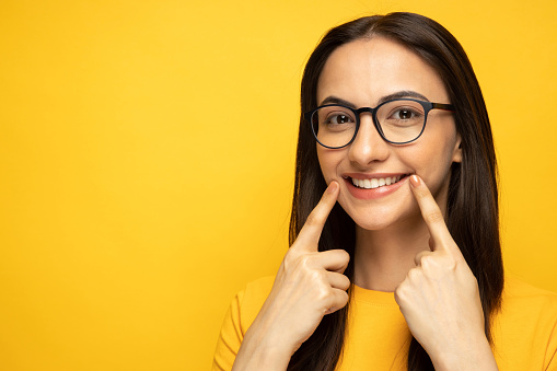 Photo of modern girl gesturing her beaming white heathy teeth with forefingers looking at camera on yellow background stock photo