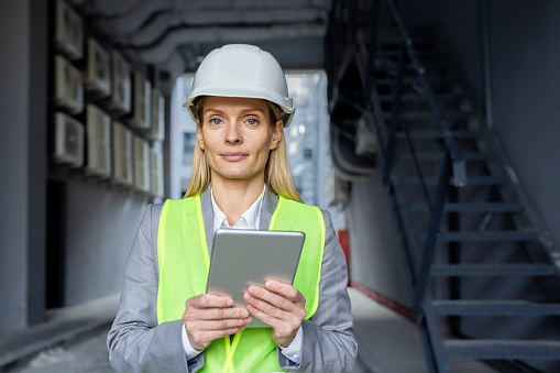 Portrait of a young female engineer, builder, architect wearing a hard hat, reflective vest and holding a tablet. He looks seriously into the camera.