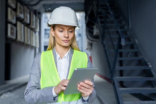 Serious confident thinking female engineer with tablet computer inspecting factory wearing hard hat and reflective vest.