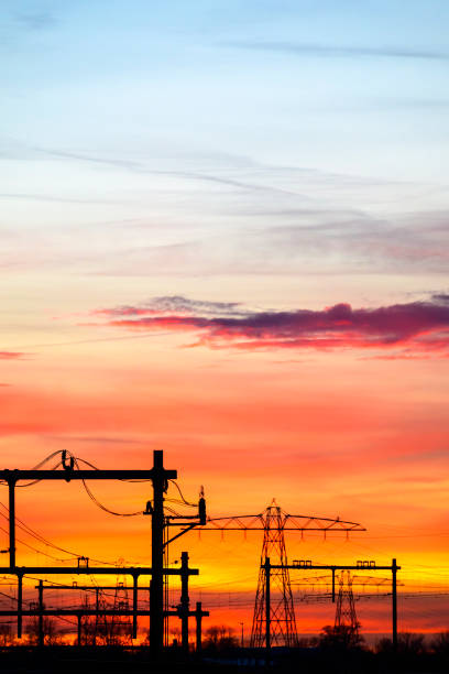 Powerline electricity pylon towers in a row next to an electrified railway track during a colorful sunset with powerlines vanishing in the distance.