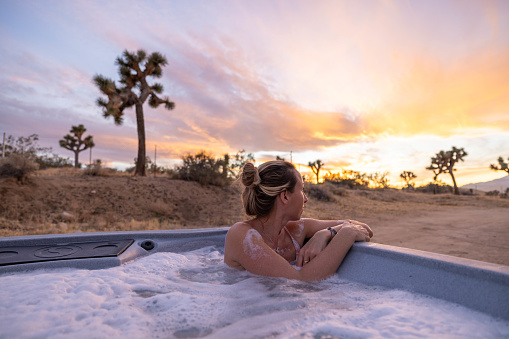 Witness a breathtaking moment as a woman immerses herself in a hot tub, surrounded by the untamed beauty of the desert. Under a spectacular sunset and a dramatic sky, nature's canvas unfolds, creating a setting of pure serenity and awe. This is a tranquil escape where relaxation harmonizes with the mystique of the desert, offering a unique and unforgettable experience.