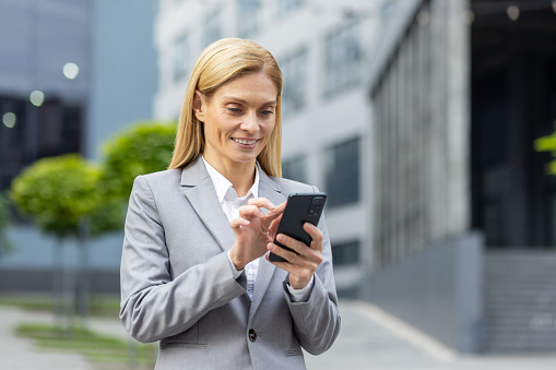 Successful woman boss walks outside office building outdoors, in business suit, businesswoman uses app on phone, smiles contentedly reads internet pages, browses news, writes text message, call.