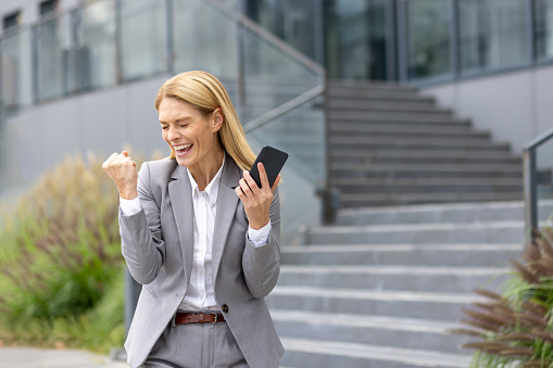 Successful mature joyful businesswoman received online notification message with good results achievement on phone, woman boss uses app on smartphone, walks outside office building in business suit.