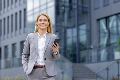 Portrait of a young businesswoman in a suit walking outside an office center, holding a phone and hand in her pocket, smiling and looking at the camera.
