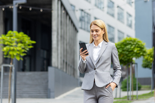 Successful woman boss walks outside office building outdoors, in business suit, businesswoman uses app on phone, smiles contentedly reads internet pages, browses news, writes text message, call.