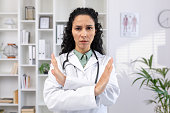 Close-up portrait of young serious hispanic female doctor standing in hospital office in white coat and crossed arms showing no sign to camera