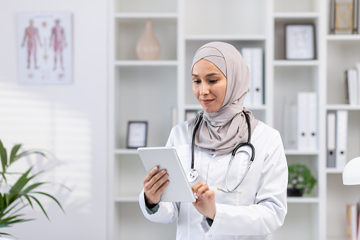Muslim woman in white medical coat standing inside medical office of clinic, female doctor using tablet computer thinking and concentrating, concentrated woman at work in hospita.l