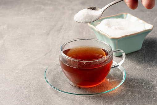 Glass cup of tea and a teaspoon with sugar in hand above the cup.