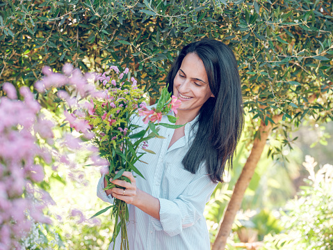 Satisfied lady with long black hair in white shirt standing in garden with blooming flowers in sunlight