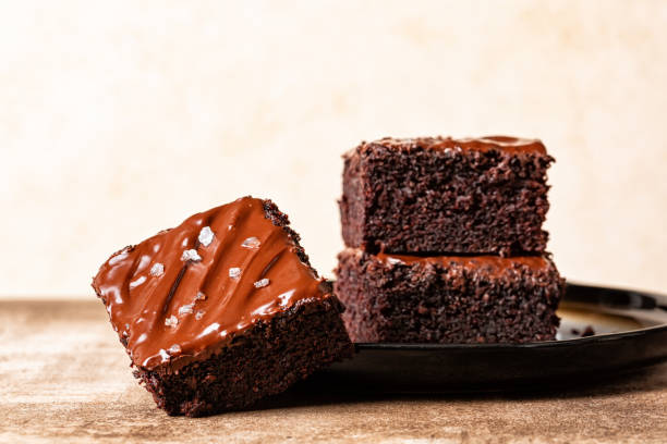 Stack of Cutted square peaces of dark chocolate moist buttermilk sheet cake with chocolate butter icing and salt, homemade sweet dessert, beige background.