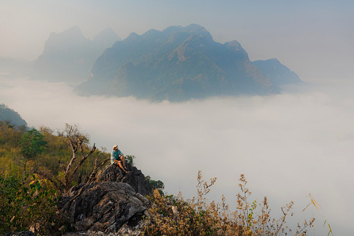Serene view of man sitting on the mountain peak and looking at fog in the valley