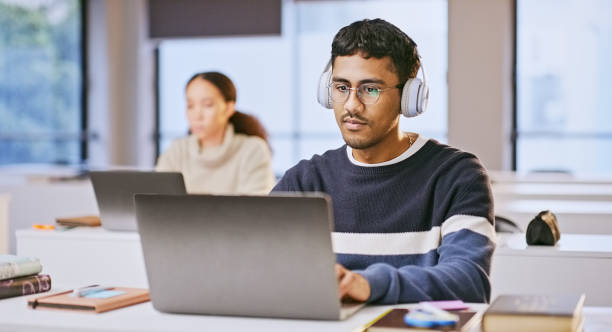 istock Laptop, headphones and student elearning in university, typing online and research assignment information. Computer, audio and man at college for education, studying knowledge and listening to music 1969854515