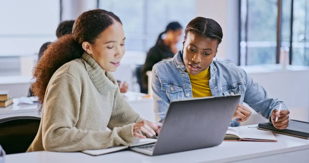 istock College, women and teamwork in classroom with laptop for elearning, reading and writing study notes. Students, diversity and friends with technology, knowledge and notebook for education or research 1969855089