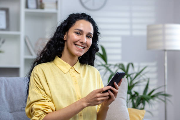 Portrait of a young beautiful hispanic woman at home, smiling and looking at the camera, holding a phone in her hands, using an application on a smartphone, browsing the Internet, typing a message