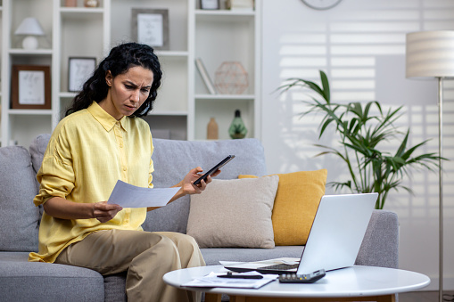 Dissatisfied and disappointed woman behind paper work sitting on sofa at home, Hispanic woman holding utility bills, creditor checks, paying upset using phone app.