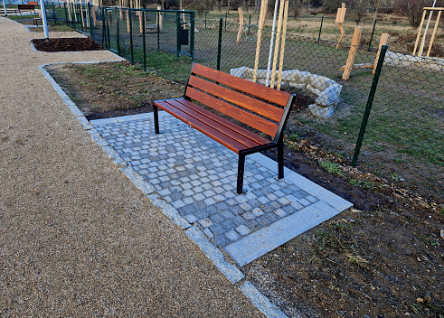 wooden modern park bench with a metal relief structure on one leg stands in the park in rest areas and on paths made of a small surface of beige gravel, lawn, perennial flower bed, wire fence, sand, threshed