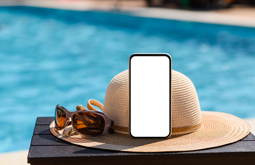 Phone with blank white display on a straw hat near the swimming pool. Holiday concept in hotels