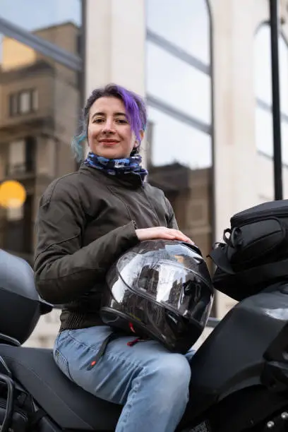 Woman smiling, sitting on her motorbike, holding her helmet, looking at the camera, low angle view.