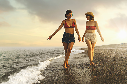 Two caucasian women in shorts and bikini tops walking on the shore, dipping their feet in the water at sunset, with sun rays behind them. One woman wears a straw hat. Both are smiling.