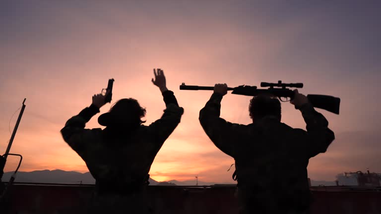 Military background of moving silhouettes of people in uniform with weapons.Soldiers waving their raised arms with rifles and pistols against the backdrop of sunset.Gestures celebrating