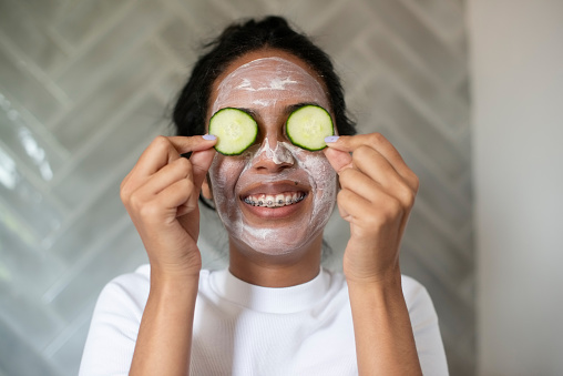 Happy woman, face mask and cucumber in bathroom for health, wellness or facial treatment at home. Young female person smile with organic vegetables in skincare for anti aging, grooming or hygiene